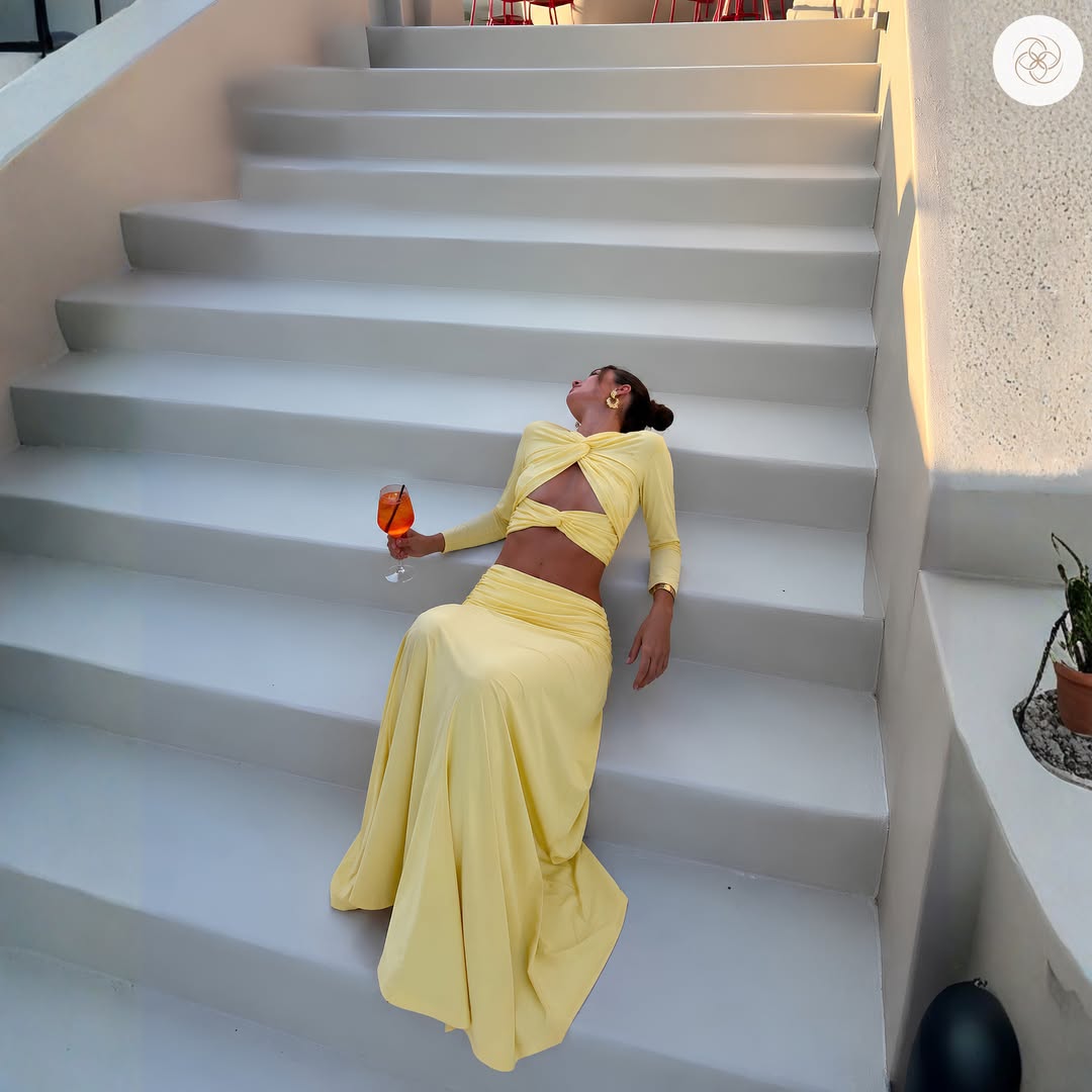 A woman reclines on white outdoor stairs wearing a pale yellow two-piece outfit with a long-sleeve crop top, front cutout detail, and matching skirt. She holds an orange drink in one hand, with a textured wall and small plant visible beside the stairs.